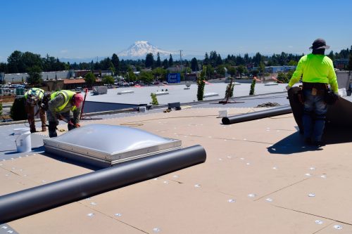 Three construction workers install materials on a flat rooftop under a clear sky, with buildings, trees, and a mountain visible in the background.