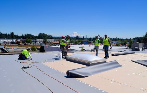 Four construction workers in safety vests install roofing material on a flat roof under a clear blue sky, with tools and equipment spread nearby.