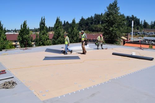 Three construction workers in safety gear install roofing material on a flat commercial rooftop on a clear day, with trees and buildings in the background.