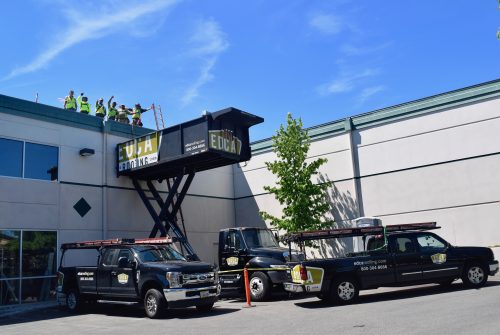 A group of workers stands on a rooftop beside a raised platform labeled "EDCA." Three black company trucks are parked below near a modern building under a clear blue sky.