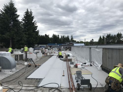 Workers in safety vests install or repair a flat commercial building roof, surrounded by equipment, tools, and materials under an overcast sky.