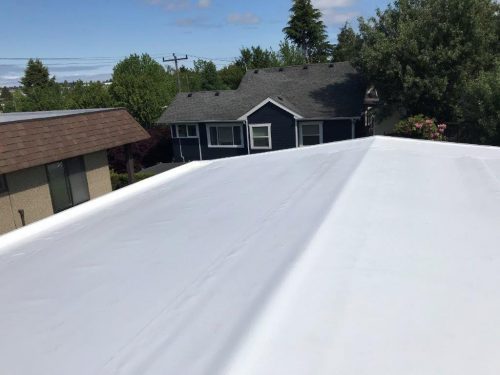 A white flat roof is shown in the foreground, with houses, trees, and a partly cloudy sky in the background.