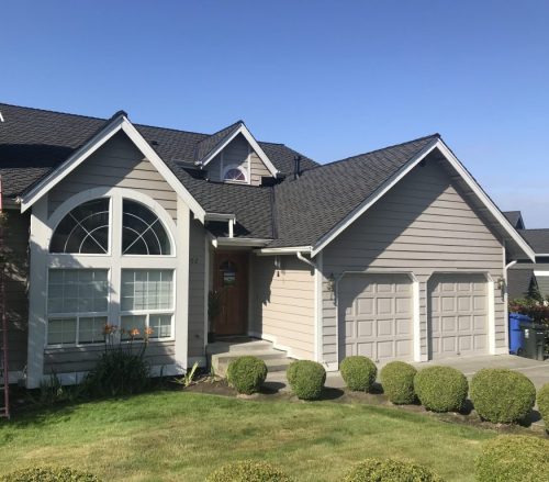 Two-story suburban house with beige siding, black roof, double garage, arched front window, wooden door, and a neatly trimmed lawn with bushes.