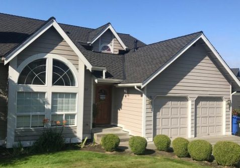 A two-story suburban house with beige siding, a large arched front window, double garage doors, and neatly trimmed bushes in the front yard.