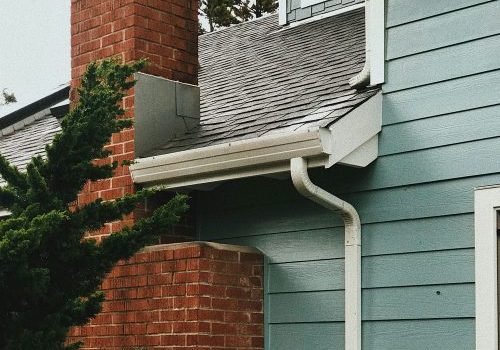 Close-up of a house exterior showing a red brick chimney, blue siding, white gutter system, and part of a sloped gray shingled roof with a tree in the foreground.